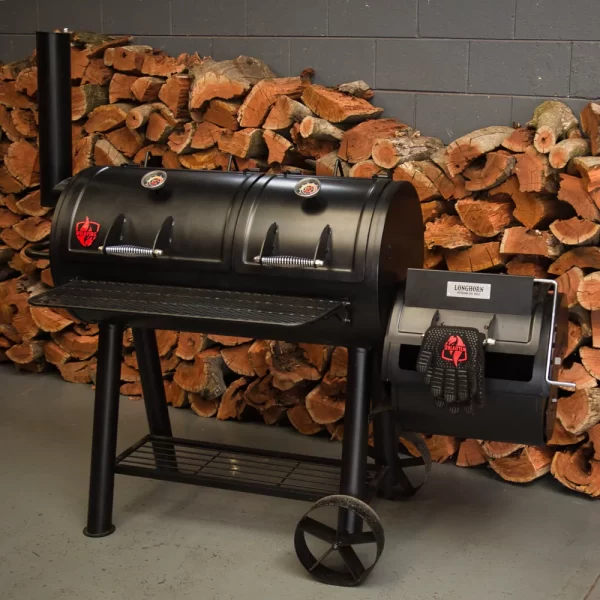 Коптильня Longhorn Offset Smoker на фоне сложенных дров — готова к приготовлению барбекю. На боковой топке висят защитные перчатки.